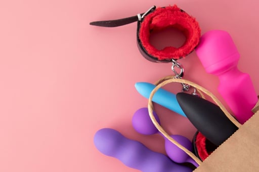 Varios juguetes sexuales asoman desde una bolsa de papel sobre un fondo rosa.