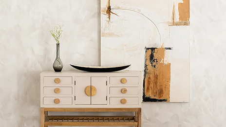 Jarrón de cristal alargado y bandeja decorativa sobre un mueble de color crema y patas de madera. Detrás se encuentra un cuadro colgado de la pared.