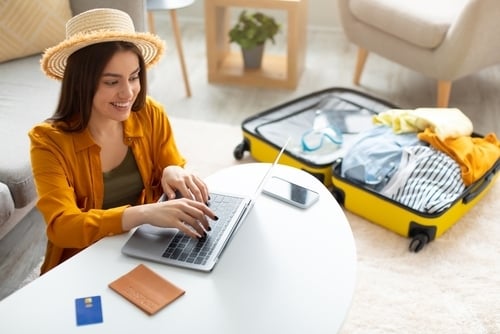Una mujer reserva unas vacaciones en su ordenador portátil. También está haciendo la maleta.